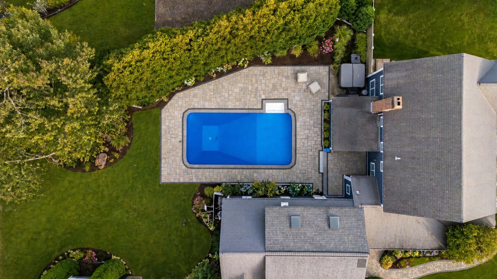 Overhead view of a completed Mayo project: a rectangular pool set into a paver terrace, bordered by planting beds and lawn, alongside a shingled house.