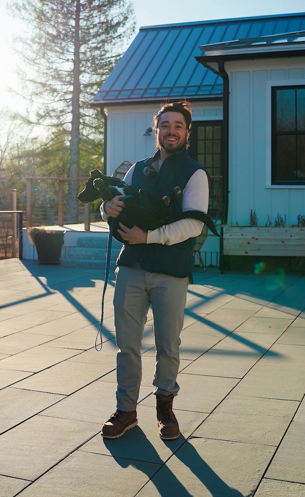 Evan Maione on a finished stone terrace in low sun, holding a dog, with a white farmhouse behind him.